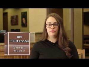 A woman wearing glasses stands in front of a sign advertising Bri Richardson, nightguards.
