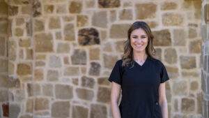 A woman in a black scrub shirt standing in front of a stone wall, practicing myofunctional therapy.